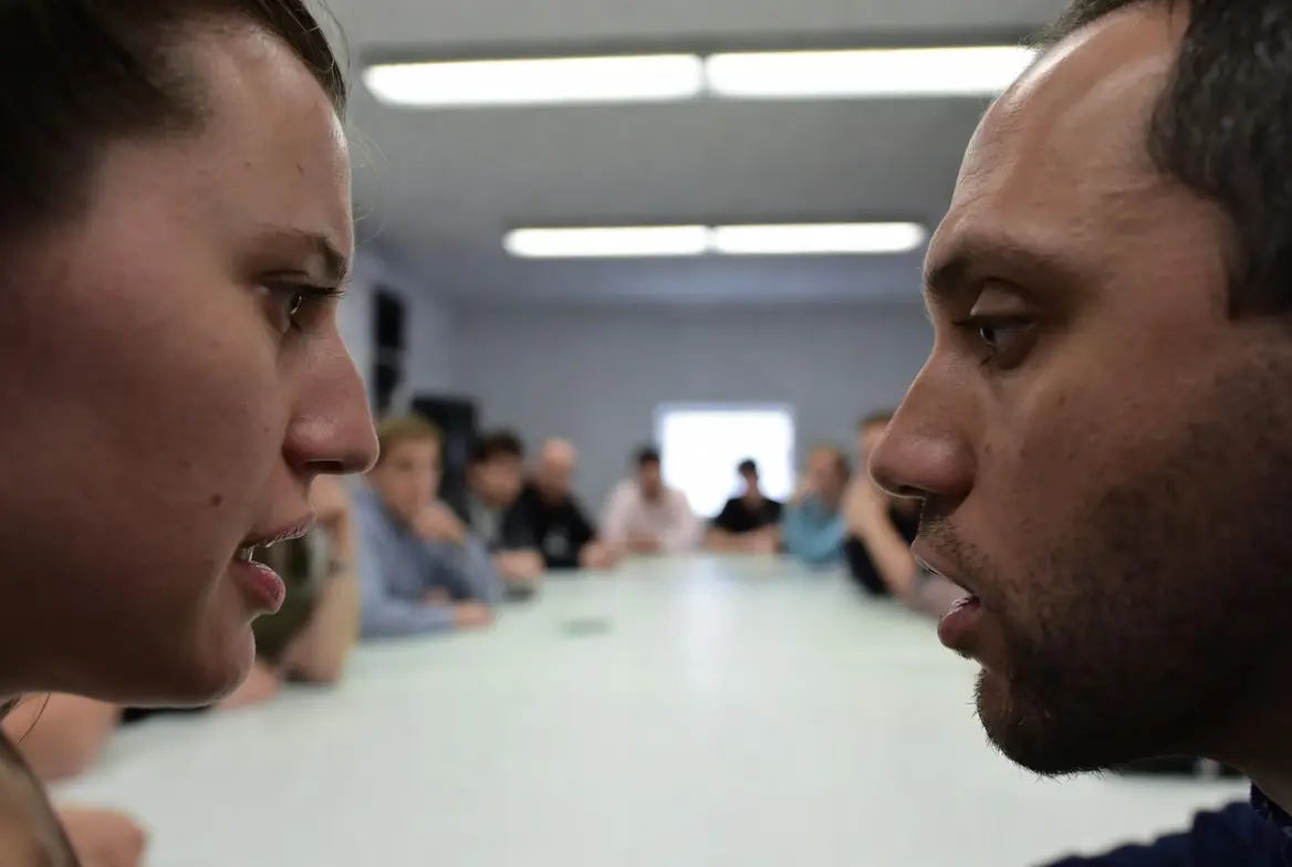 To personer sitter ansikt til ansikt ved et møtebord i et konferanserom og ser direkte på hverandre med alvorlige uttrykk, mens flere kolleger sitter rundt bordet i bakgrunnen.